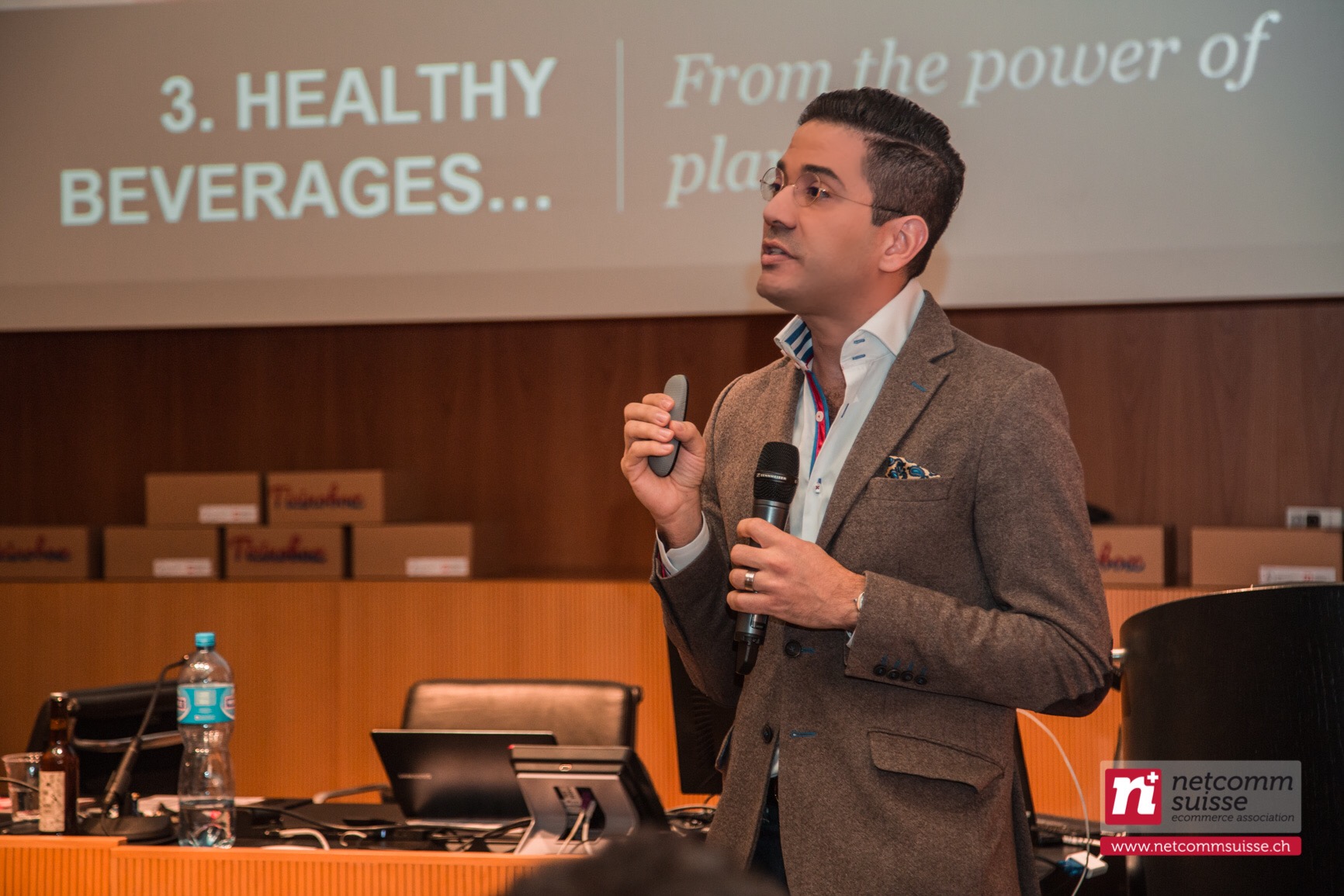 José Namén presenting with a microphone at a Netcomm Suisse ecommerce event, slides projected behind him.