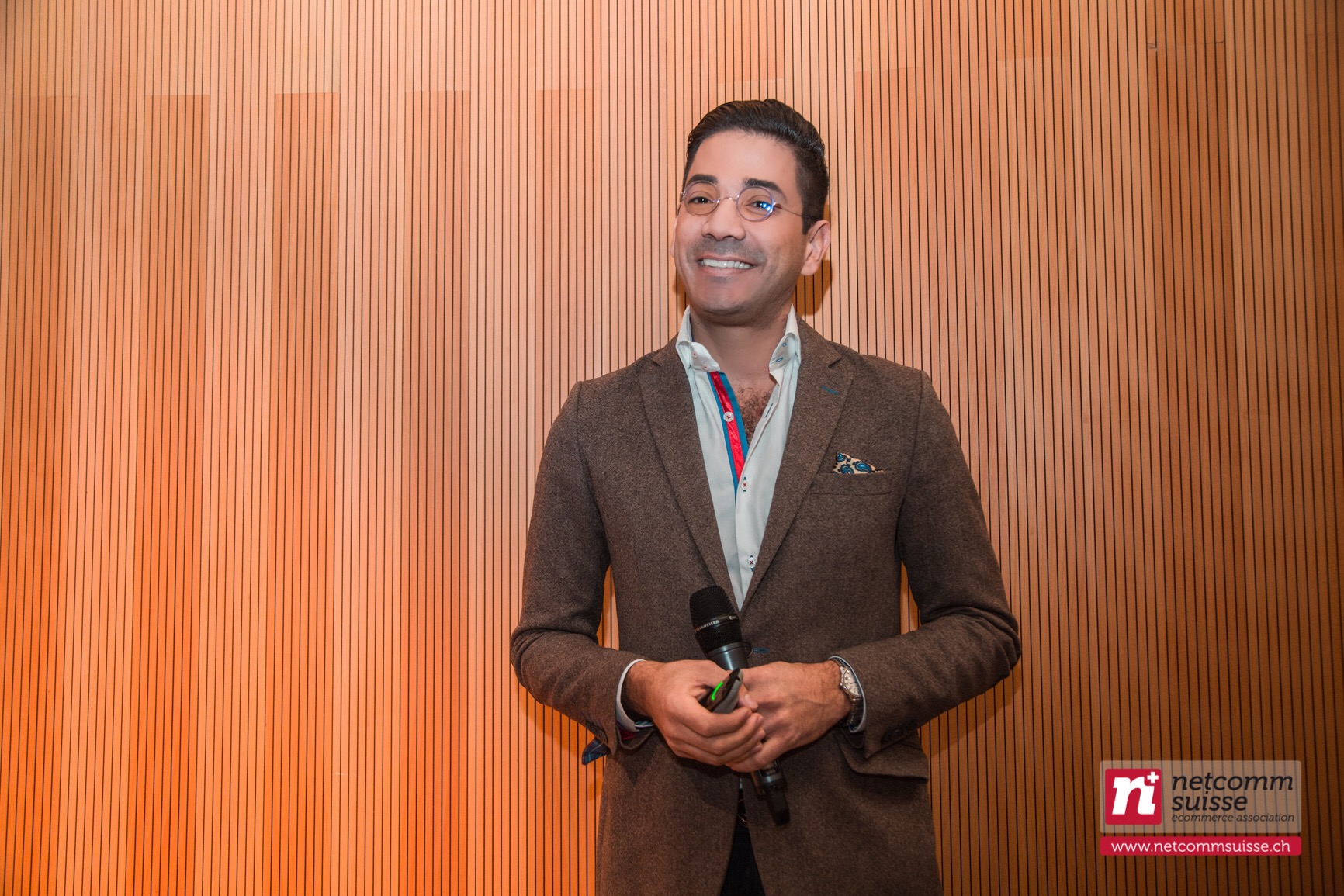José Namén smiling, holding a microphone, brown blazer against a wooden slat wall at a Netcomm Suisse event.